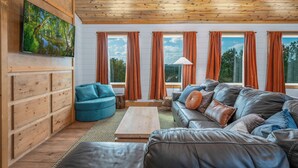 Ein gemütliches Wohnzimmer mit Holzboden, einem großen Flachbildfernseher, der an einer Holzvertäfelung angebracht ist, einem grauen Ledersofa, einem türkisfarbenen Sessel und einem Holztisch.