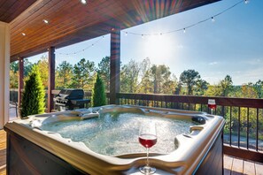 Ein Whirlpool mit einem Glas Rotwein auf einer Holzterrasse, beleuchtet mit Lichterketten, mit Blick auf ein bewaldetes Gebiet.