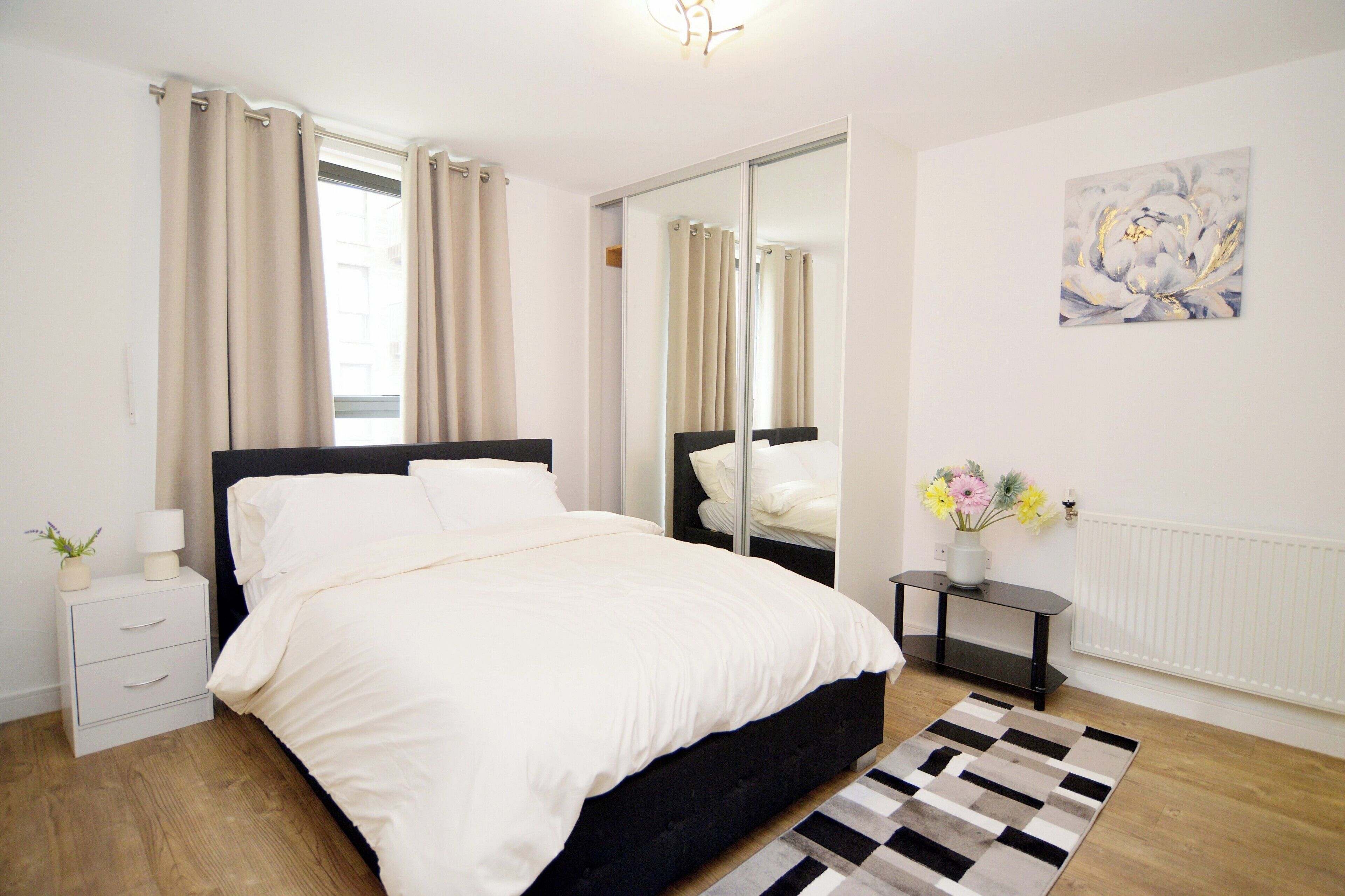 Une chambre moderne avec un lit recouvert de tissu noir, une table de chevet avec une lampe, une commode, un miroir et un radiateur.