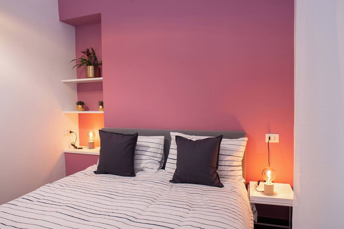 A bedroom with a bed, two pillows, a nightstand, a lamp, and a shelf with a plant.