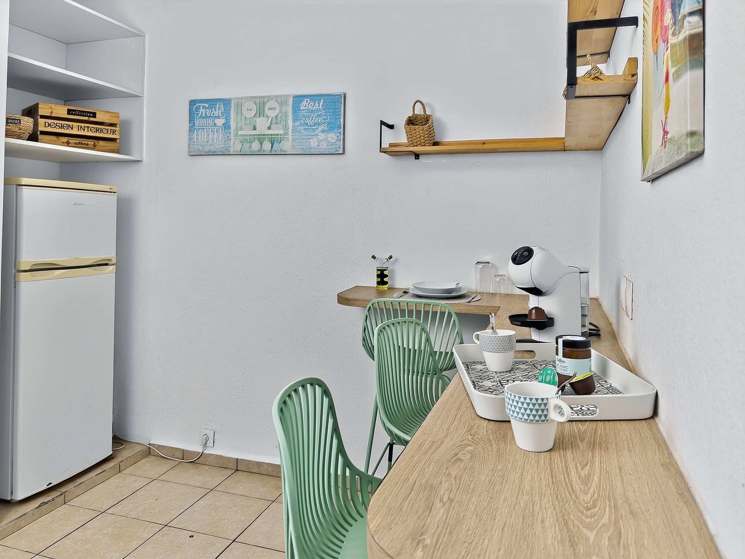 A compact kitchen with a refrigerator, a dining table with chairs, a coffee machine, and a shelf with decorative items.
