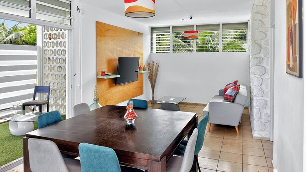 A modern dining area with a wooden table, grey and blue chairs, a TV mounted on a wooden panel, and a view of greenery outside.