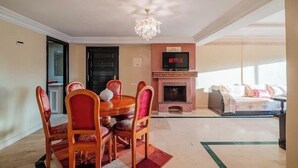 A living room with a fireplace, a dining area with a round table and chairs, a television mounted on the wall, and a bed in the background. - Luxe Appt 2 CH Piscine & Parking (Marrakech)