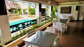 A covered outdoor seating area with tables and chairs, a pool view, and a thatched-roof structure.
