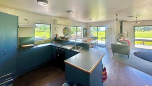 A spacious kitchen with a large island, a dining area with a table and chairs, and a living area with a sofa. There are large windows providing views of the outdoors. - Family Home on Golden Bay Farm — Peaceful, Kid-Friendly Stay (Puramāhoi)
