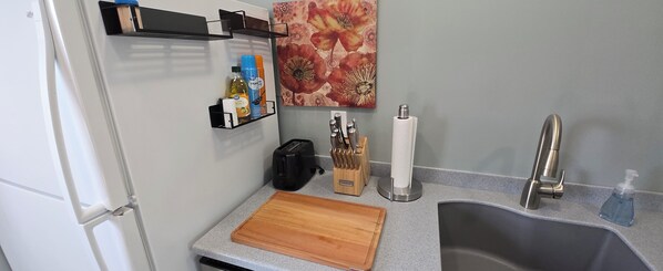 A kitchen with a wooden cutting board, a sink, a toaster, a knife block, and a flower painting on the wall.