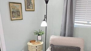 Una habitación con pared azul claro, un sillón color beige con cojín gris, alfombra estampada, una mesita con una planta y dos cuadros enmarcados.