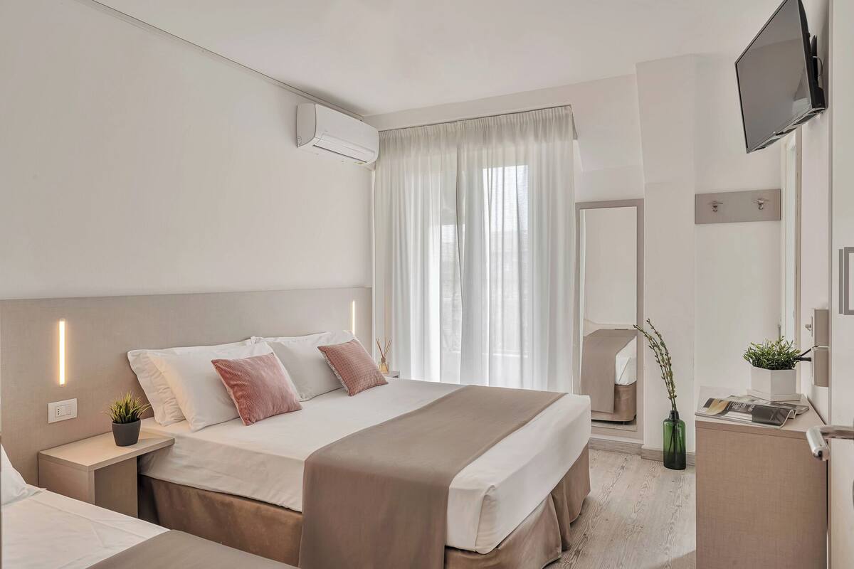 A hotel room with a bed, bedside tables, a TV, a mirror, and a window with curtains.
