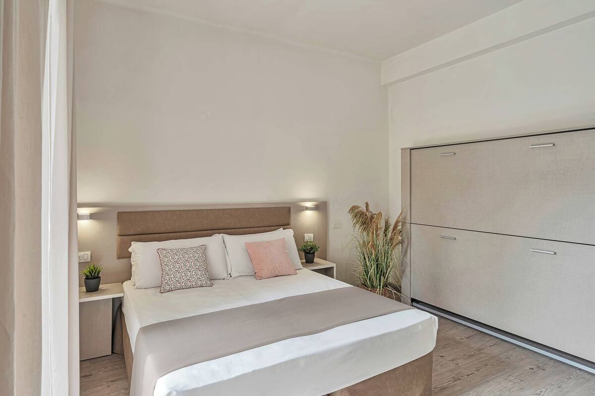 A bedroom with a bed, bedside tables, a nightstand, a wardrobe, and a potted plant.