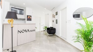 Uma área de recepção com balcão identificado como "NOVA SUITE", um abajur preto com design de elefante, uma poltrona de veludo preto e um vaso com planta.