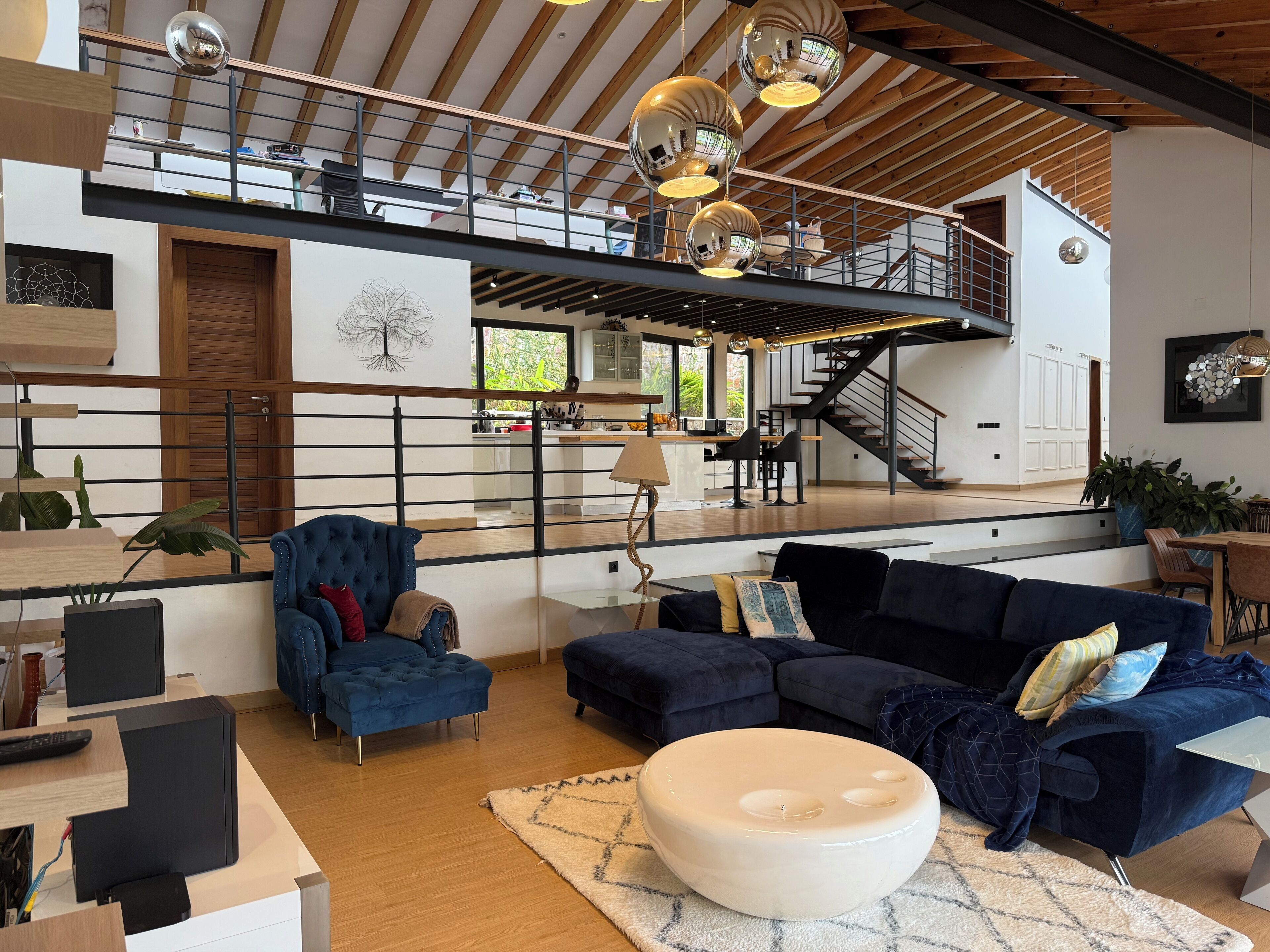 A spacious living area with a high ceiling, a staircase, and a kitchen in the background.