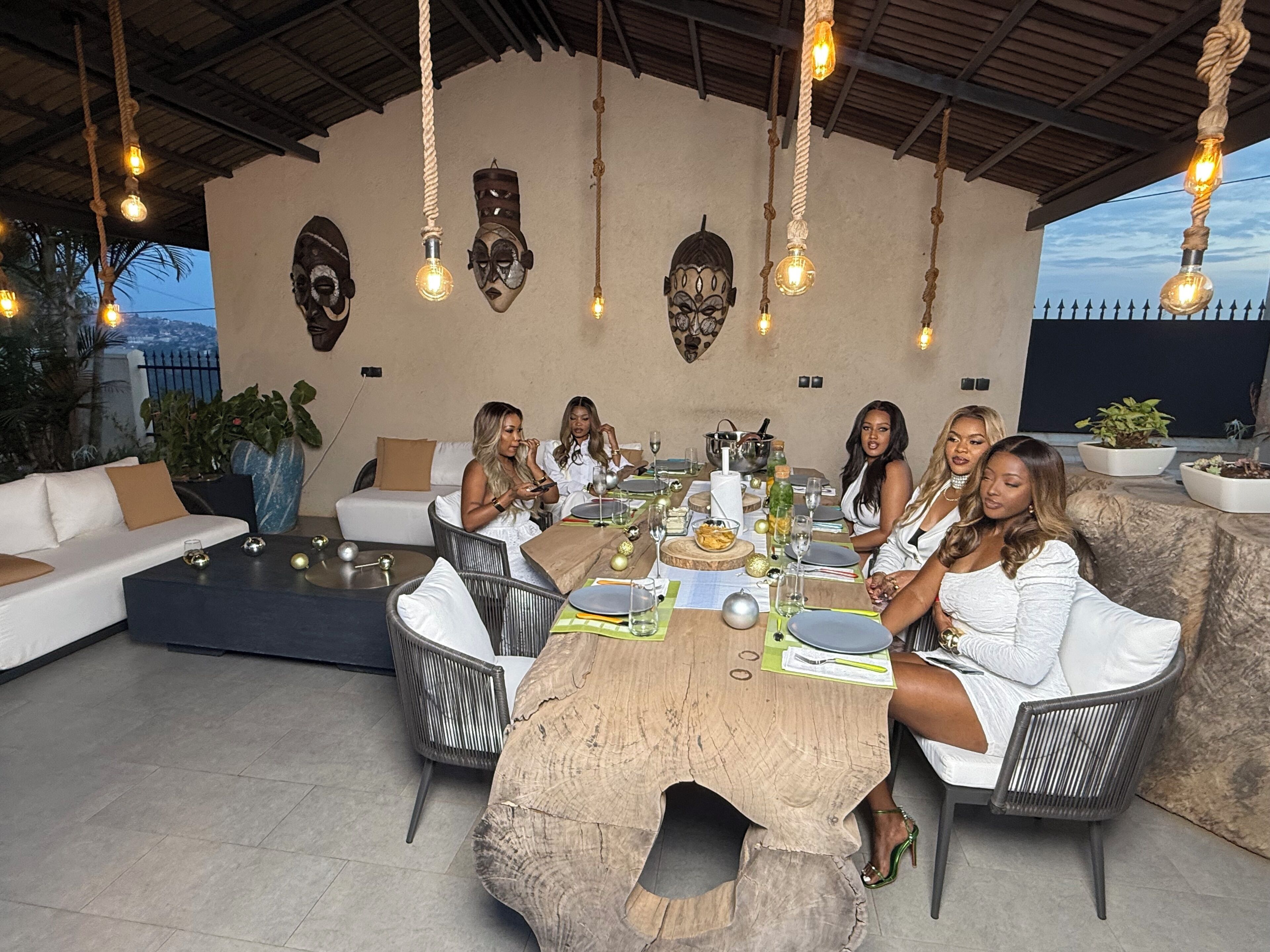 A group of people seated around a wooden table with a rustic design, under a covered area with hanging light fixtures.