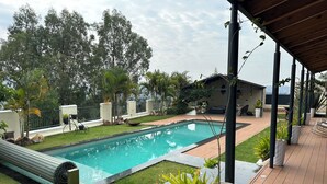 Ein rechteckiger Pool mit einer umlaufenden Holzterrasse, einer überdachten Veranda und verschiedenen Kübelpflanzen.