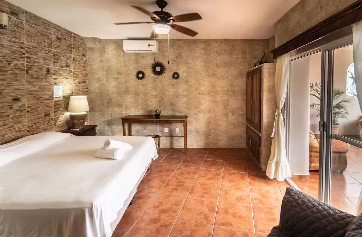 Une chambre d’hôtel avec un lit, un bureau, un ventilateur de plafond et une vue sur l’extérieur, visible à travers une porte vitrée.