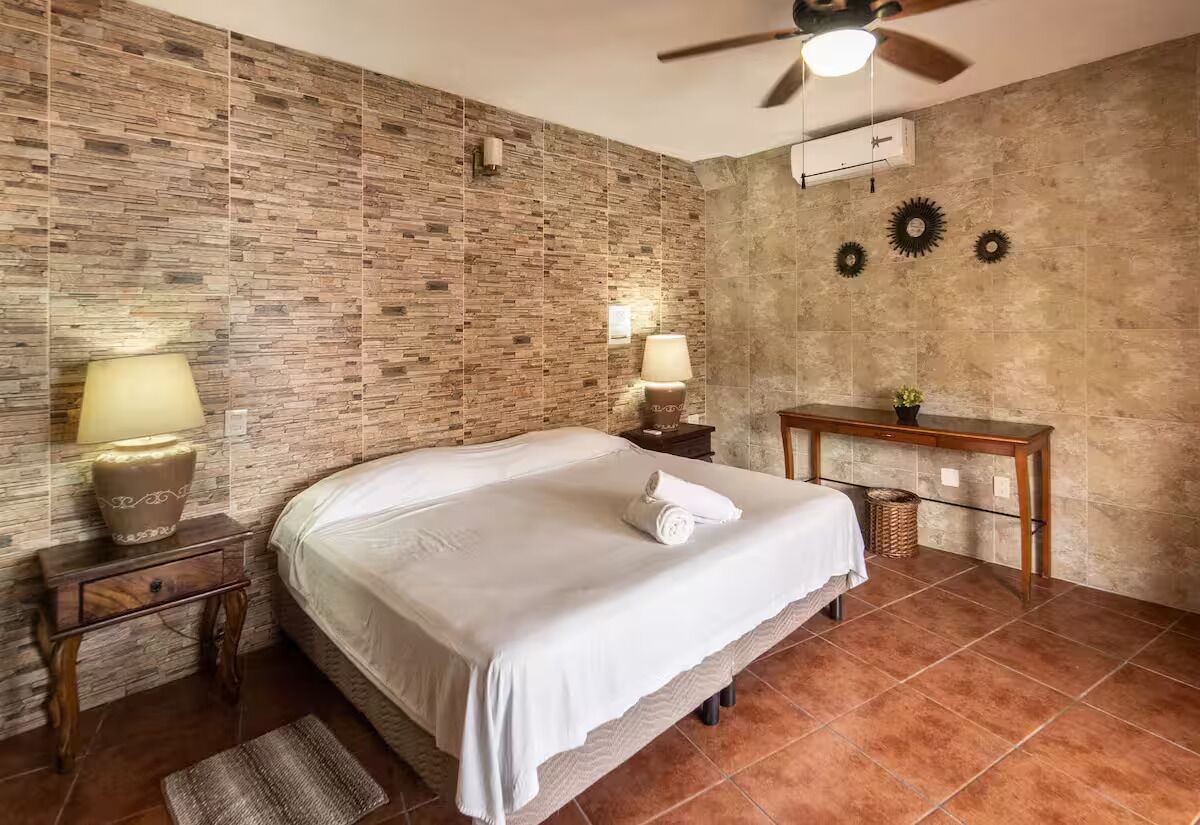 A bedroom with a brick-patterned wall, a bed with white bedding, a ceiling fan, and a small table with a lamp.