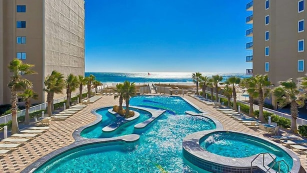 A resort pool area with multiple swimming pools, palm trees, and a view of the ocean. - Crystal Tower 1802: 2BR Condo (Gulf Shores)