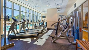 A gym with treadmills, ellipticals, and weight machines, large windows, and a view of palm trees. - Crystal Tower 1802: 2BR Condo (Gulf Shores)