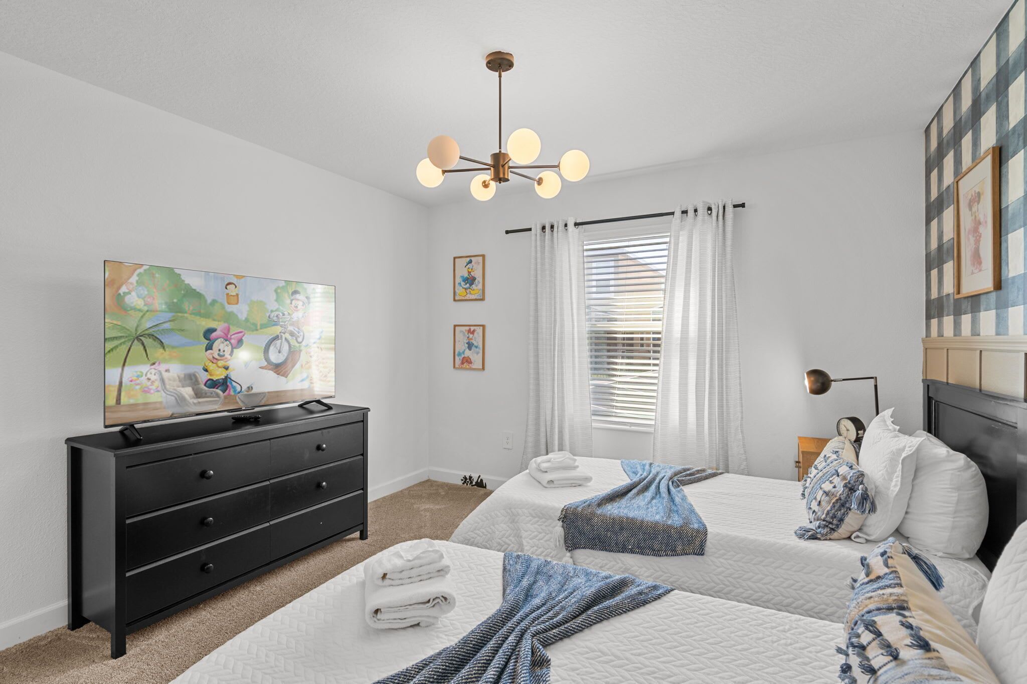 A hotel room with two beds, a flat-screen TV, a black dresser, a ceiling light fixture, and a window with blinds.