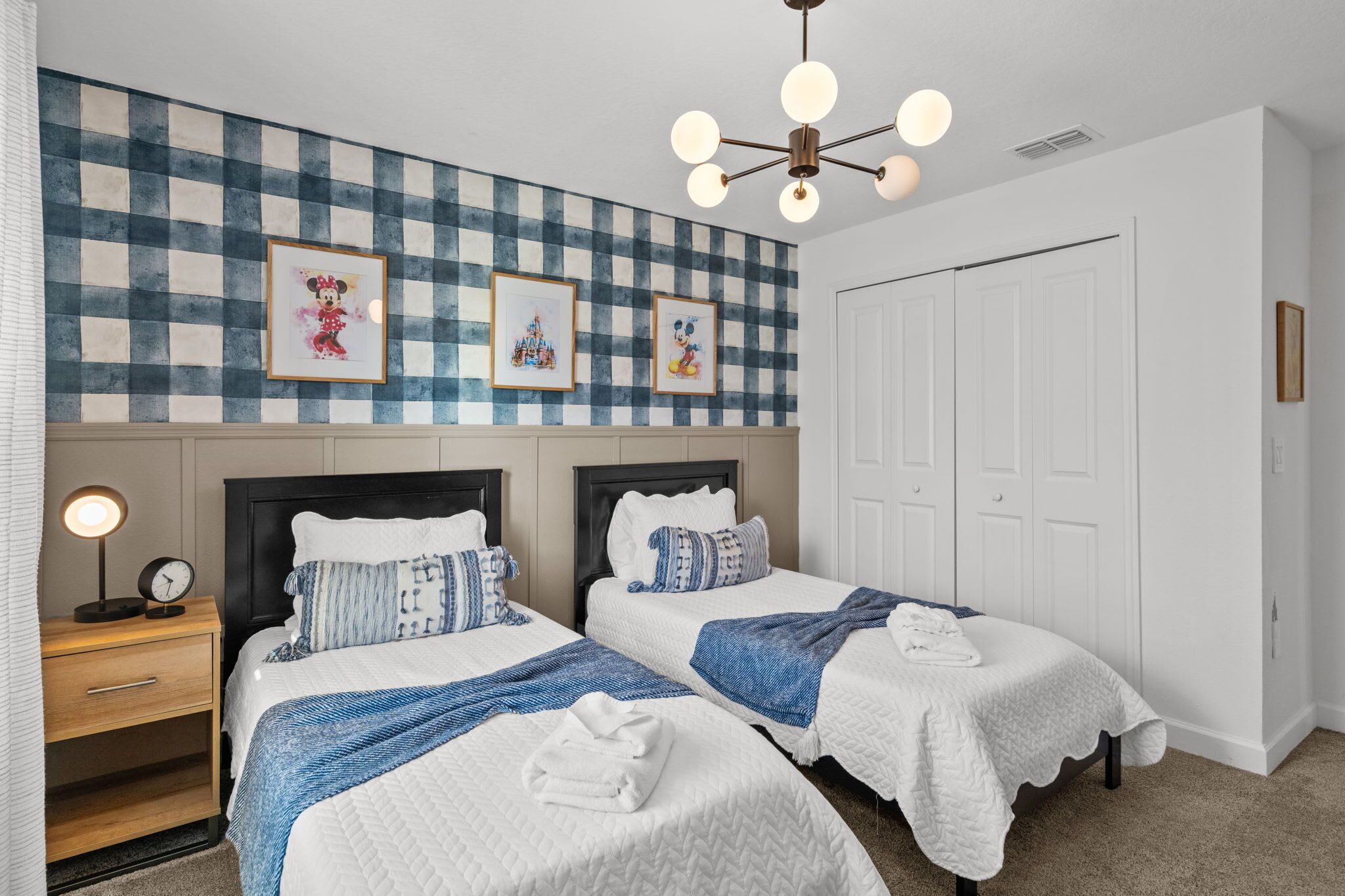 A hotel room with two beds, a checkered wall, framed pictures, a bedside lamp, and a white door.