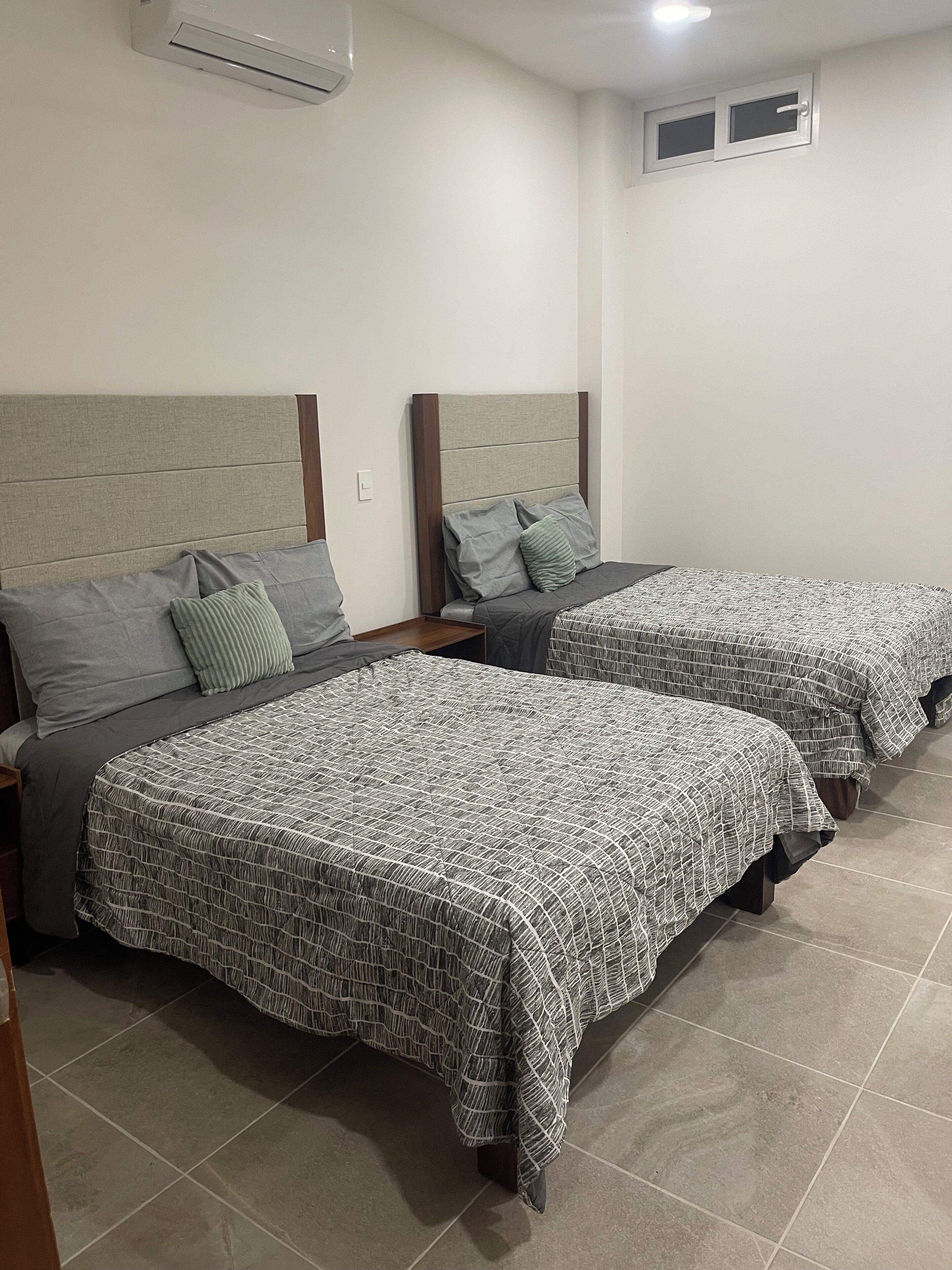 A hotel room with two beds, each with a headboard, grey bedding, and a small bedside table.