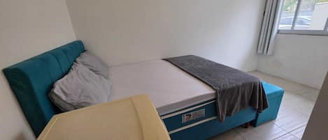 Una habitación pequeña con cama individual, sofá azul, una mesita y una ventana con cortinas.