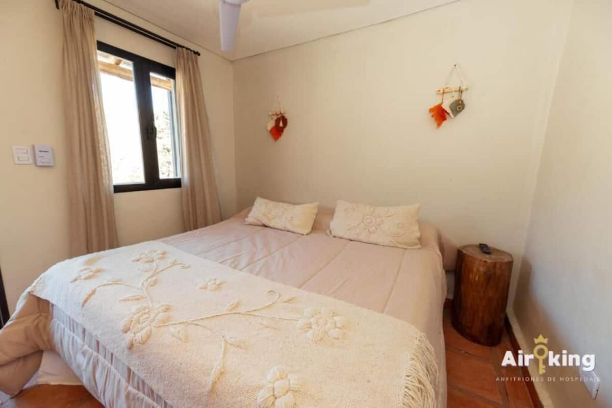 Un dormitorio con una cama grande, cortinas color beige, un taburete de madera y una lámpara fijada a la pared.