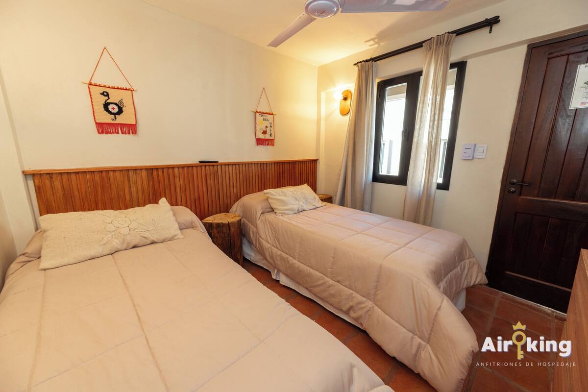 Habitación de hotel con dos camas, cabecero de madera, ventilador de techo, ventana con cortinas y una puerta con un cartel.