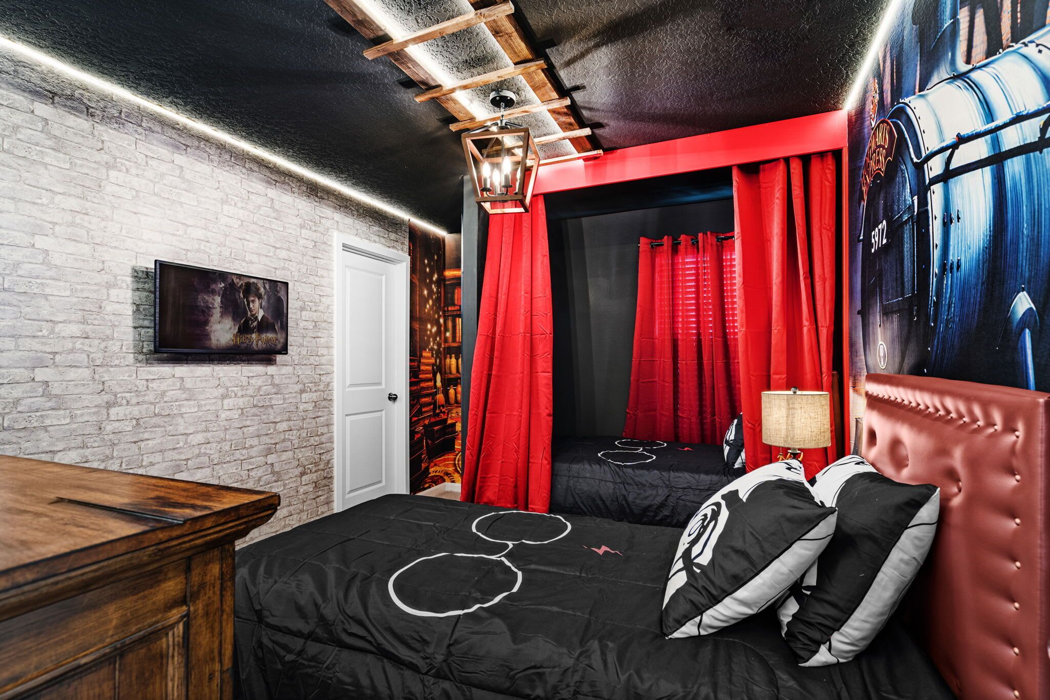 Ein Zimmer mit Backsteinwand, einem Holztisch, einem Bett mit schwarzer Bettwäsche und weißen kreisförmigen Mustern, roten Vorhängen und einem Wandbild eines Zuges.