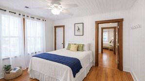 Une chambre à coucher avec un lit, un plancher en bois, un ventilateur de plafond et une porte donnant sur une autre pièce.
