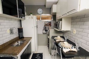 A compact kitchen with white cabinets, a granite countertop, a clock, and a dining area set with plates and glasses.