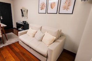A living room with a beige sofa, a small table, a mirror, and framed leaf art on the wall.