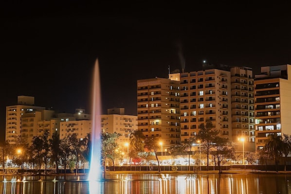A nighttime cityscape with a prominent fountain, illuminated buildings, and a calm water body reflecting the lights.