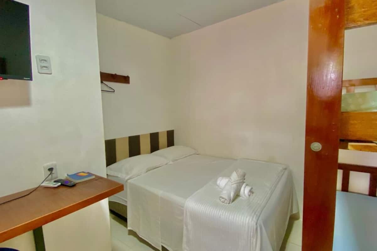 A single bed with a white blanket and a towel, a wooden desk with a chair, and a television mounted on the wall.