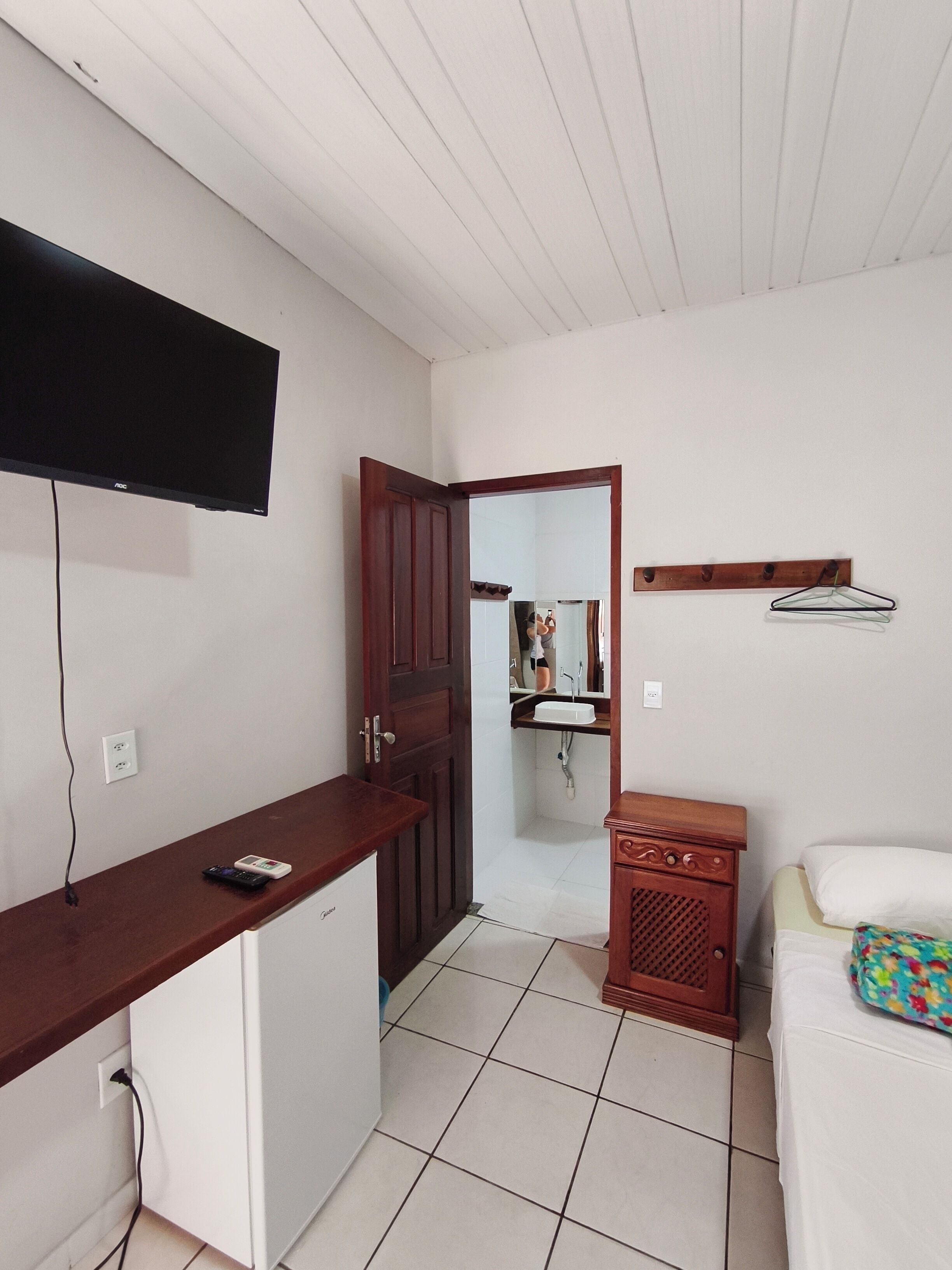 A compact hotel room with a kitchenette, a television mounted on the wall, a wooden cabinet, a small bedside table, and a bed with a colorful pillow.