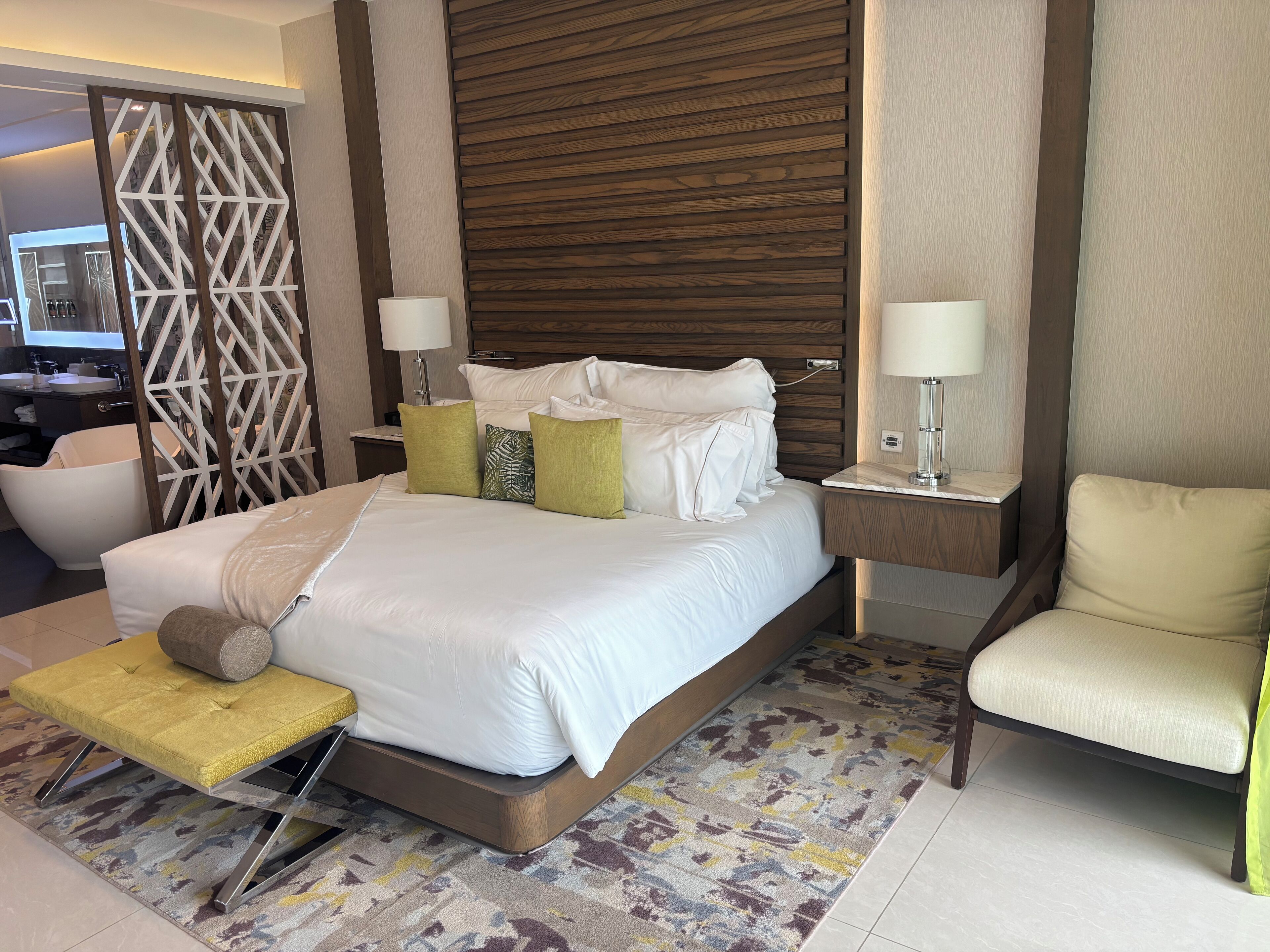 Une chambre d’hôtel moderne avec un grand lit, deux tables de chevet, une chaise, un canapé et un élément de séparation décoratif.