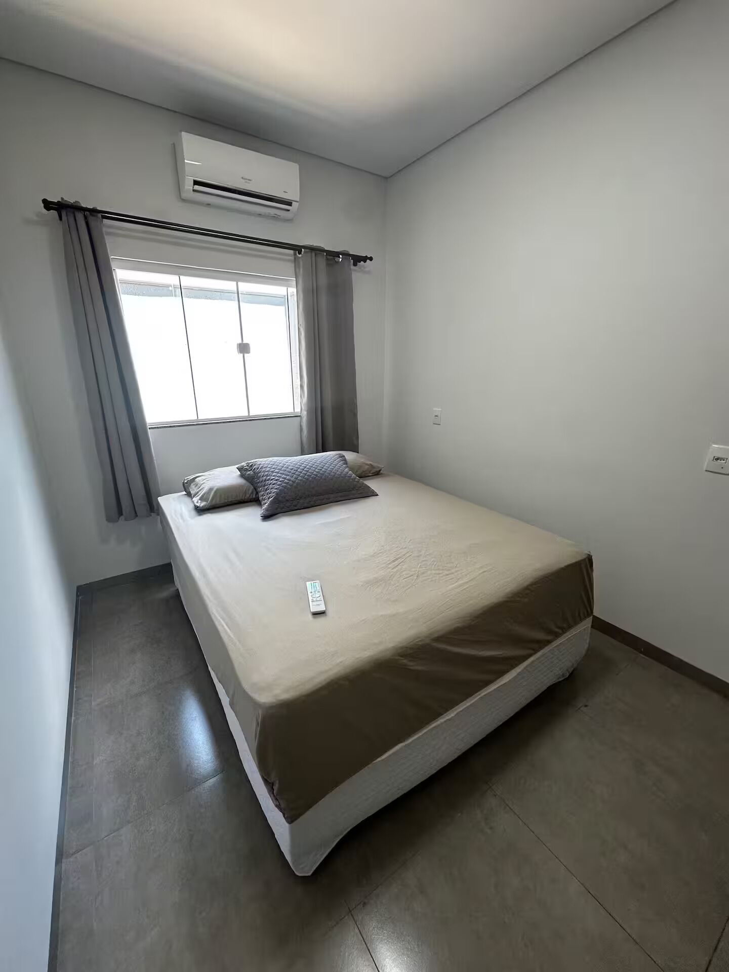 Una habitación pequeña y limpia con cama, aire acondicionado y ventana con cortinas.