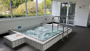 Ett bubbelbad med en metallstege, omgivet av vita kakelväggar och ett fönster med utsikt över grönskan.