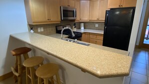 A kitchen with wooden cabinets, a black refrigerator, a microwave, a sink, and a granite countertop. There are three wooden stools under the counter. - Red Mountain Village 2 bedroom condo sleeps 6 (Rossland)
