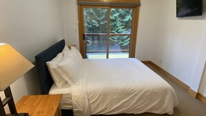 A bedroom with a bed, a nightstand, a lamp, a TV, and a window with a view of trees. - Red Mountain Village 2 bedroom condo sleeps 6 (Rossland)