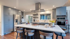A modern kitchen with grey cabinets, a central island with bar stools, stainless steel appliances, and a large window. - Retreat With Lake Access, Deck & Modern Interior Agate Peak by Avantstay (Carnelian Bay)