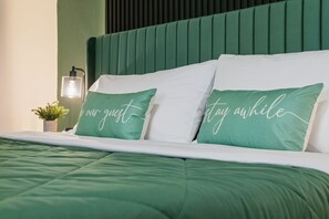 Ein ordentlich bezogenes Bett mit grüner Bettwäsche und zwei Kissen, eines mit dem Schriftzug "no our guest" und das andere mit "stay awhile".