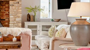 Ein gemütliches Wohnzimmer mit einem weißen Sofa, einer flauschigen Decke und einer großen Lampe. Eine Backsteinwand und ein Fenster mit Blick auf die Landschaft sind ebenfalls zu sehen.