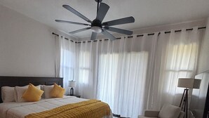 Une chambre à coucher comprenant un lit, une chaise, un bureau, un ventilateur de plafond et des rideaux.