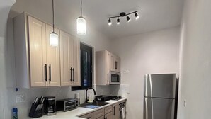 Une cuisine compacte avec des meubles blancs, un réfrigérateur en acier inoxydable, un four à micro-ondes et une cuisinière à gaz. On y trouve deux tabourets de bar et un petit îlot central avec une machine à café et un grille-pain.