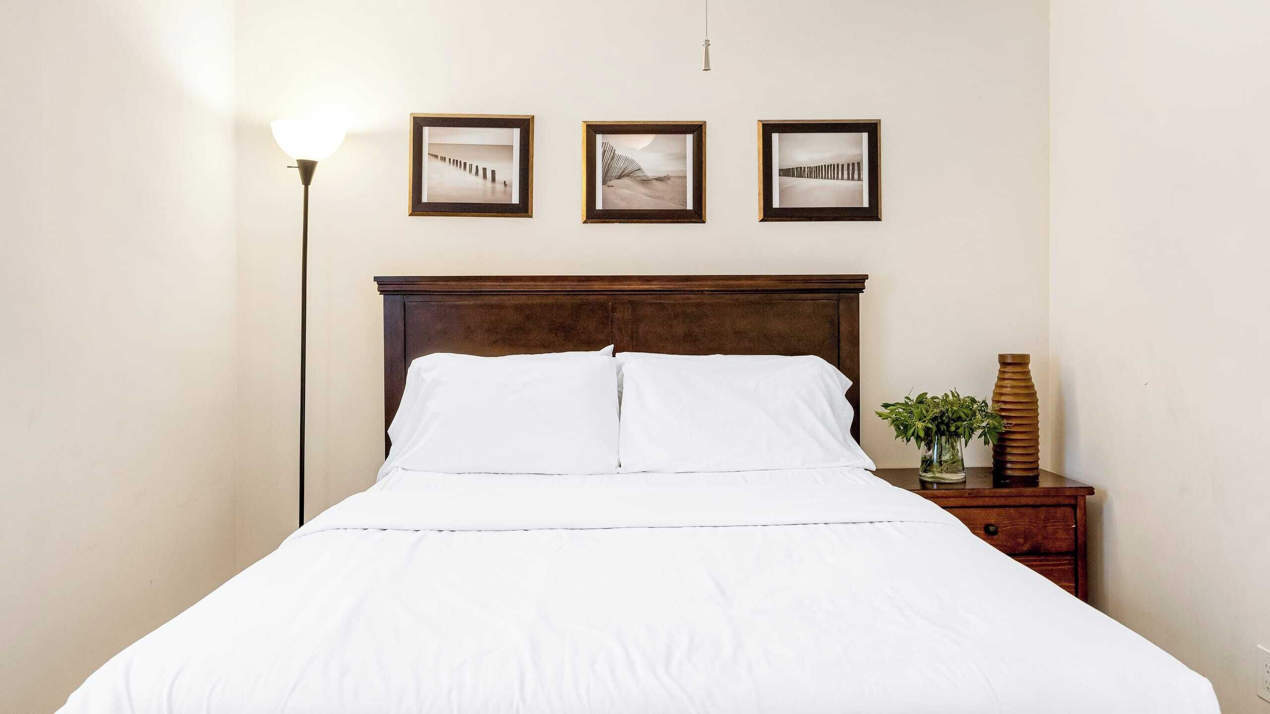 Una cama bien hecha con sábanas blancas, un cabecero de madera y tres cuadros enmarcados en la pared de arriba.