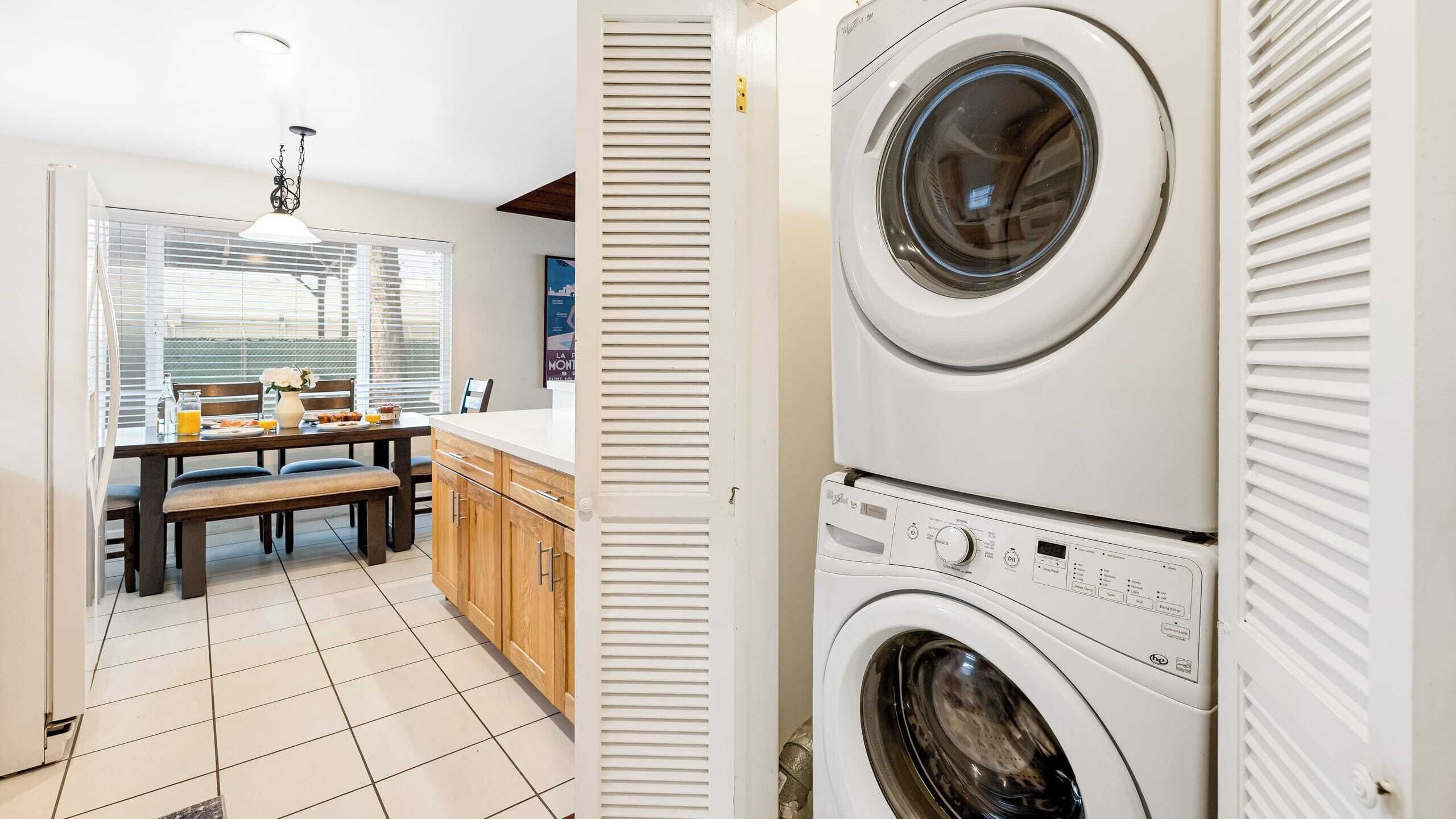 Une buanderie avec des lave-linge et des sèche-linge superposés, un coin repas avec une table et des chaises, et une fenêtre avec des stores.