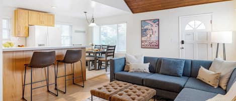 A modern kitchen with wooden cabinets, a dining area with a table and chairs, a living room with a sofa and ottoman, and a ceiling with wooden beams.