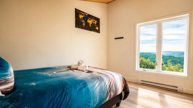 Un dormitorio con un ventanal grande, una cama con cubrecama azul, un cuadro en la pared y vistas a zonas verdes al exterior.