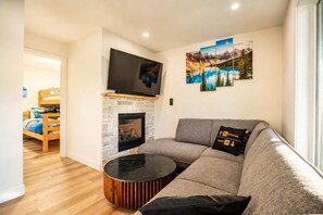 Ein Wohnzimmer mit Kamin aus Stein, einem Flachbildfernseher, einer Couch mit Sitzkissen und einem Essbereich mit Holztisch und Stühlen.