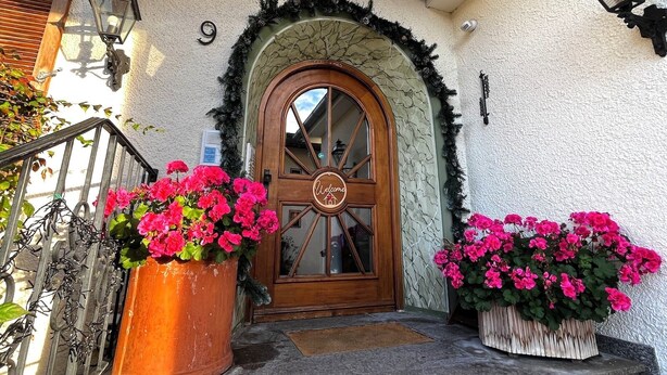 En dörr av trä med ett runt fönster och en välkomstskylt, flankerad av två stora blomkrukor.